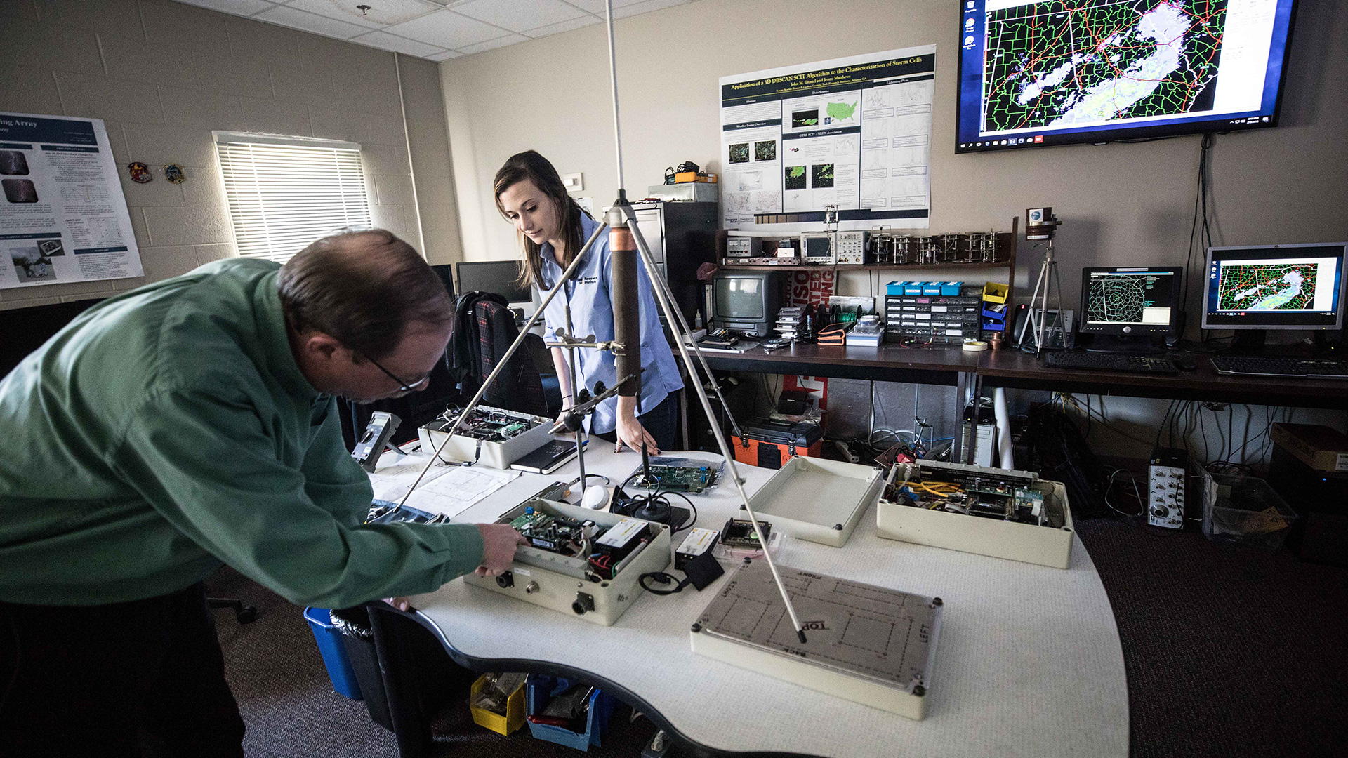 photo: a young woman and older man standing at a table containing weather instruments, in the Severe Storms Research Center Lab.