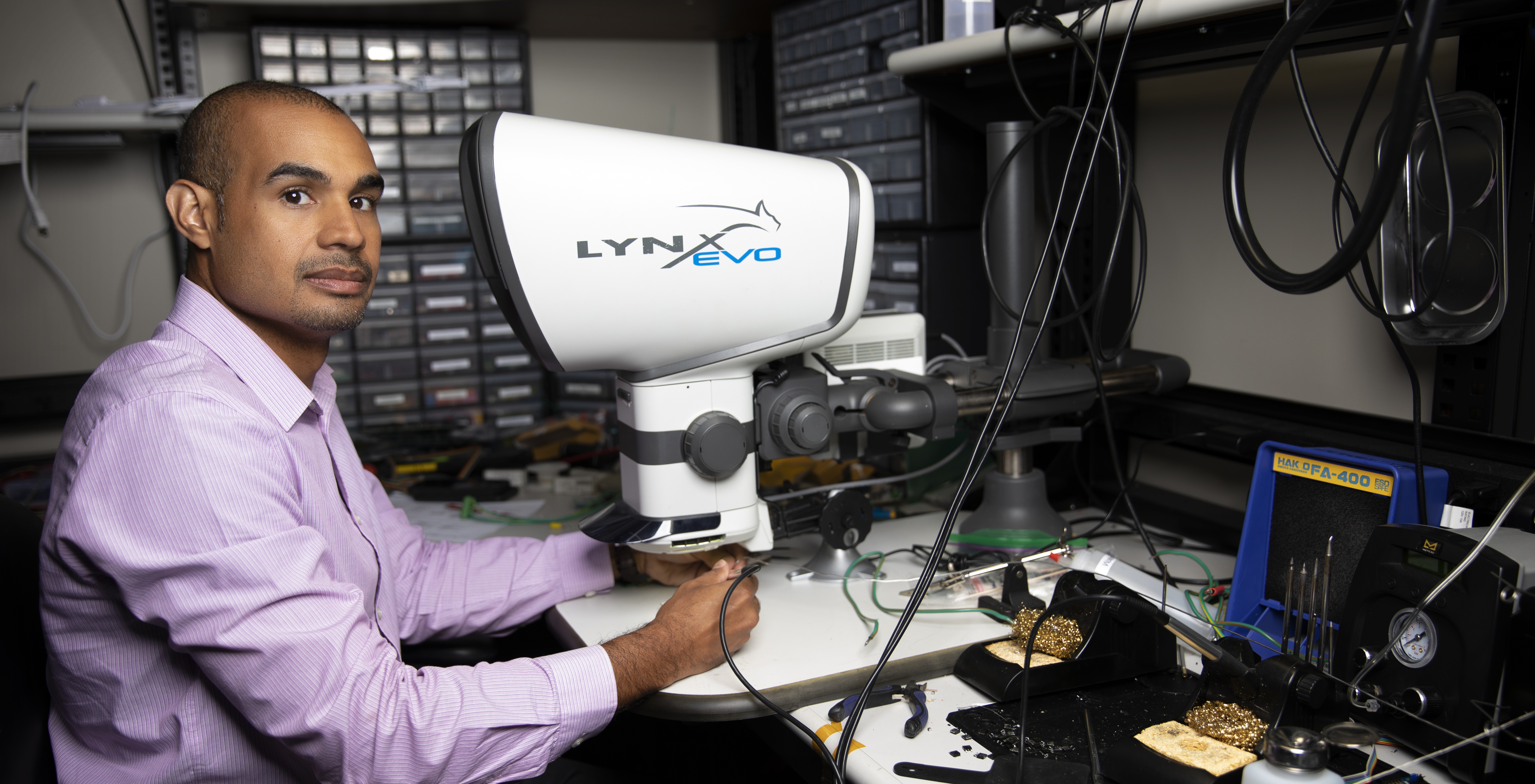 Mike Ruiz sits at a desk with a eyepiece-less microscope and various technical equipment. (Photo credit: Sean McNeil)